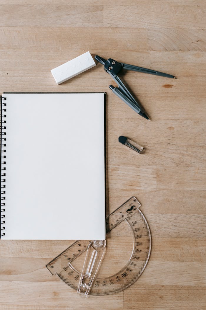 Flat lay of school supplies including notebook, compass, eraser, and protractor on wooden desk.